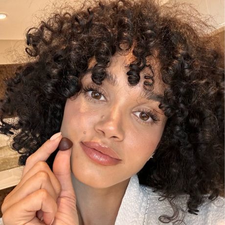 Smiling woman with curly hair holding a HAIR GUMMY by Rite – supports healthy hair growth with vitamins and plant-based nutrients
