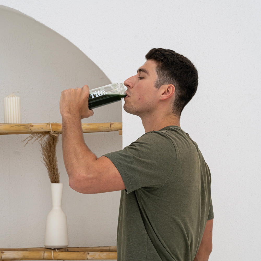 A man in a green shirt drinking a Daily Greens powder from a glass bottle labeled ""rite"" against a minimalist background with a shelf and decorative items.







