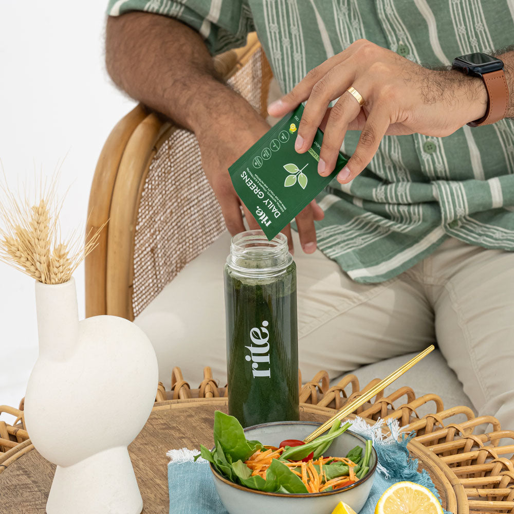 A man pours a green powder packet labeled "Rite Daily Greens" into a green shaker bottle. There is a salad and a lemon on the table.