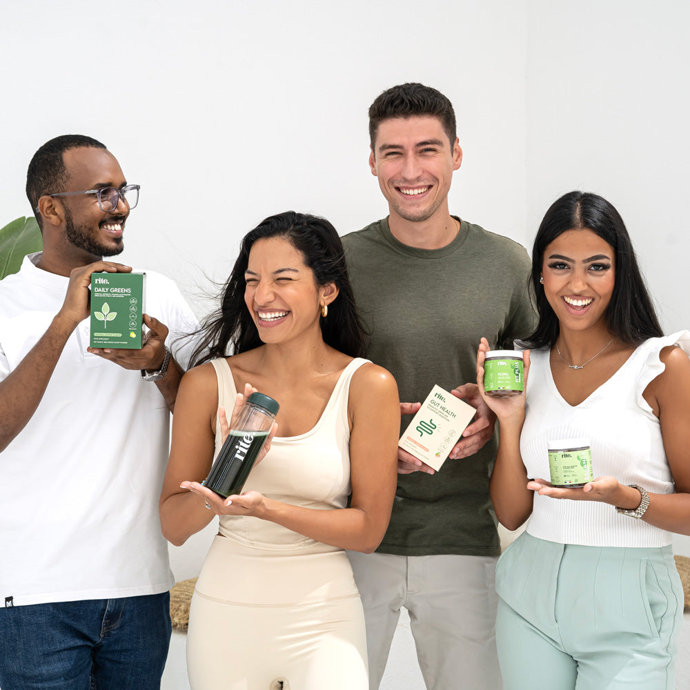 Four smiling people holding various health products, including supplements and a shaker bottle, standing together in a bright, minimalist setting.