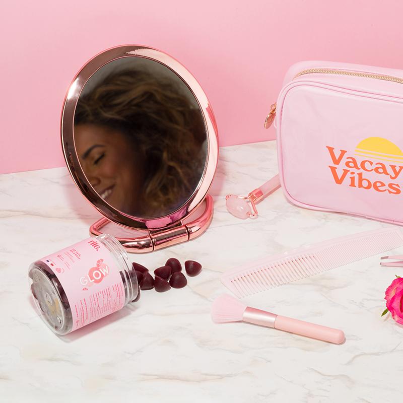 A collection of beauty products and  GLOW gummies with a reflection of a smiling woman in a round mirror.