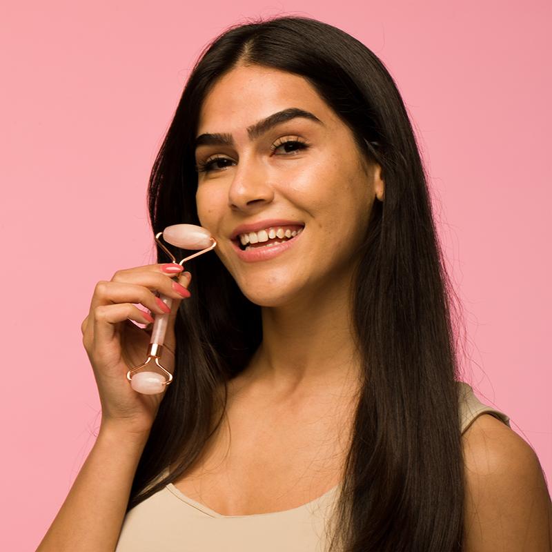A smiling woman holding a facial roller against a pink background.